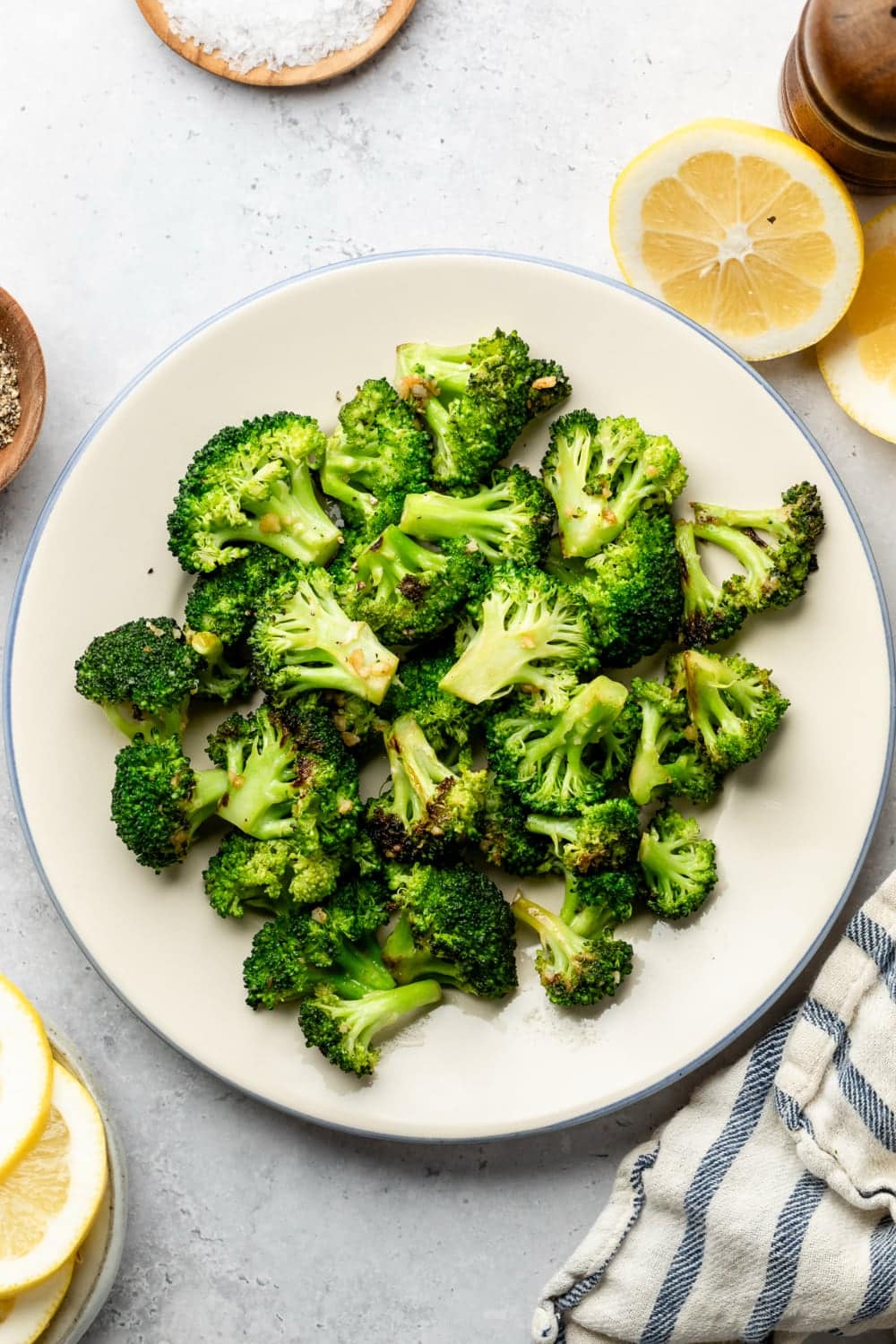 A plate with sautéed garlic butter broccoli ready for serving surrounded by lemon wedges and additional salt and pepper.