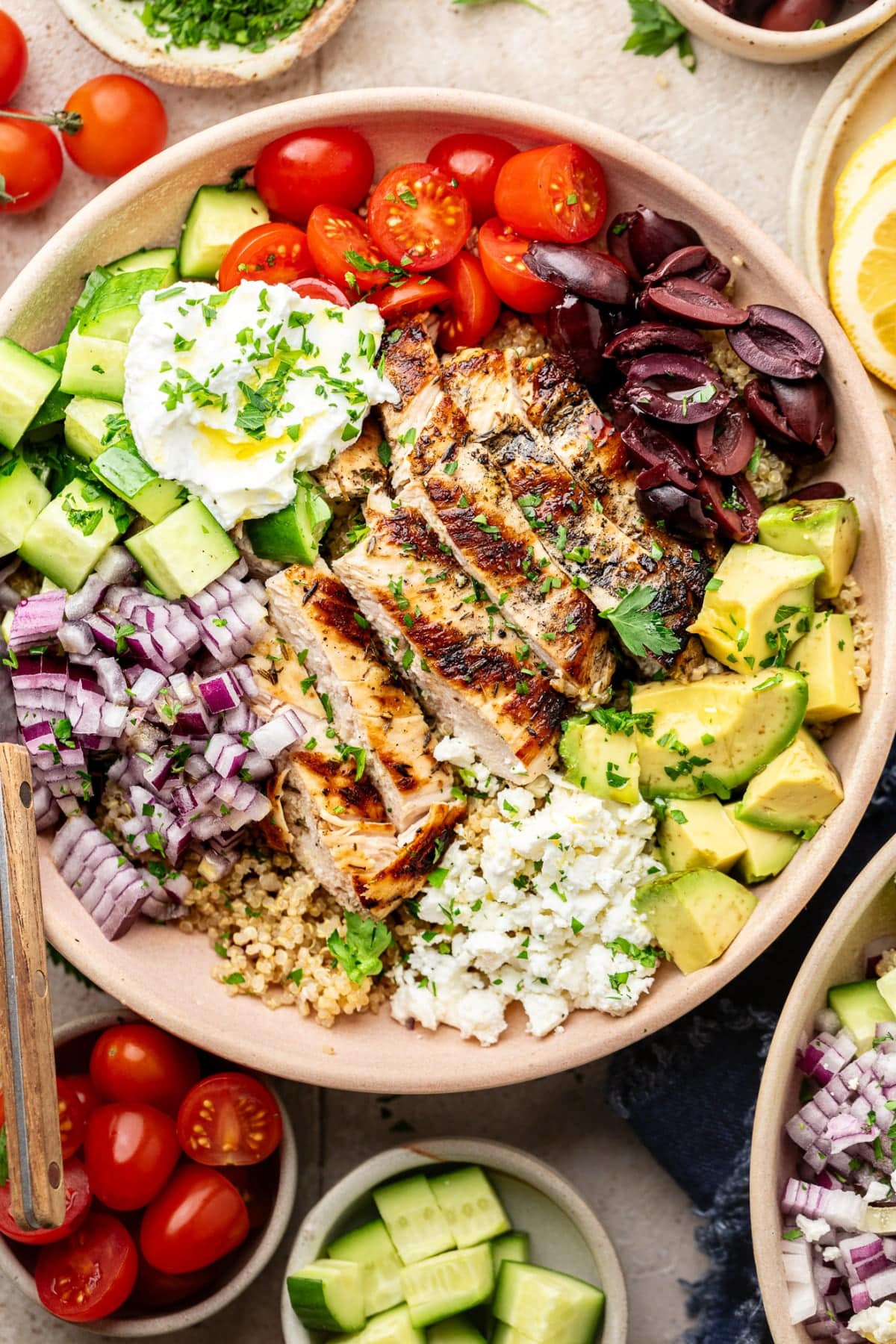 A large brown bowl with a fully assembled Greek chicken quinoa bowl topped with feta cheese, olives, and Greek yogurt. There is a fork in the bowl ready to eat.