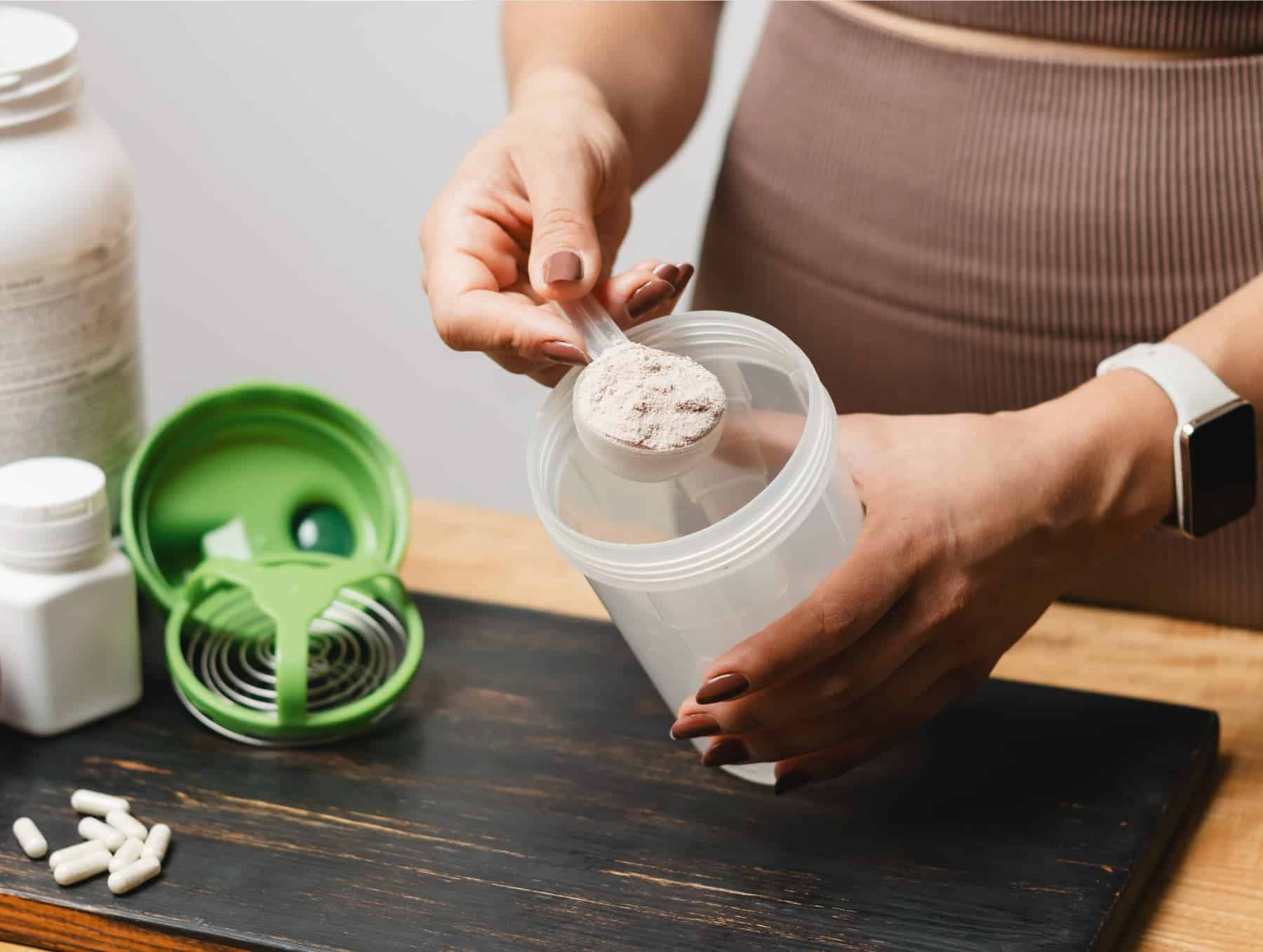 A woman scooping protein powder and creatine into a plastic shaker bottle post workout with supplements on the table in front of her.