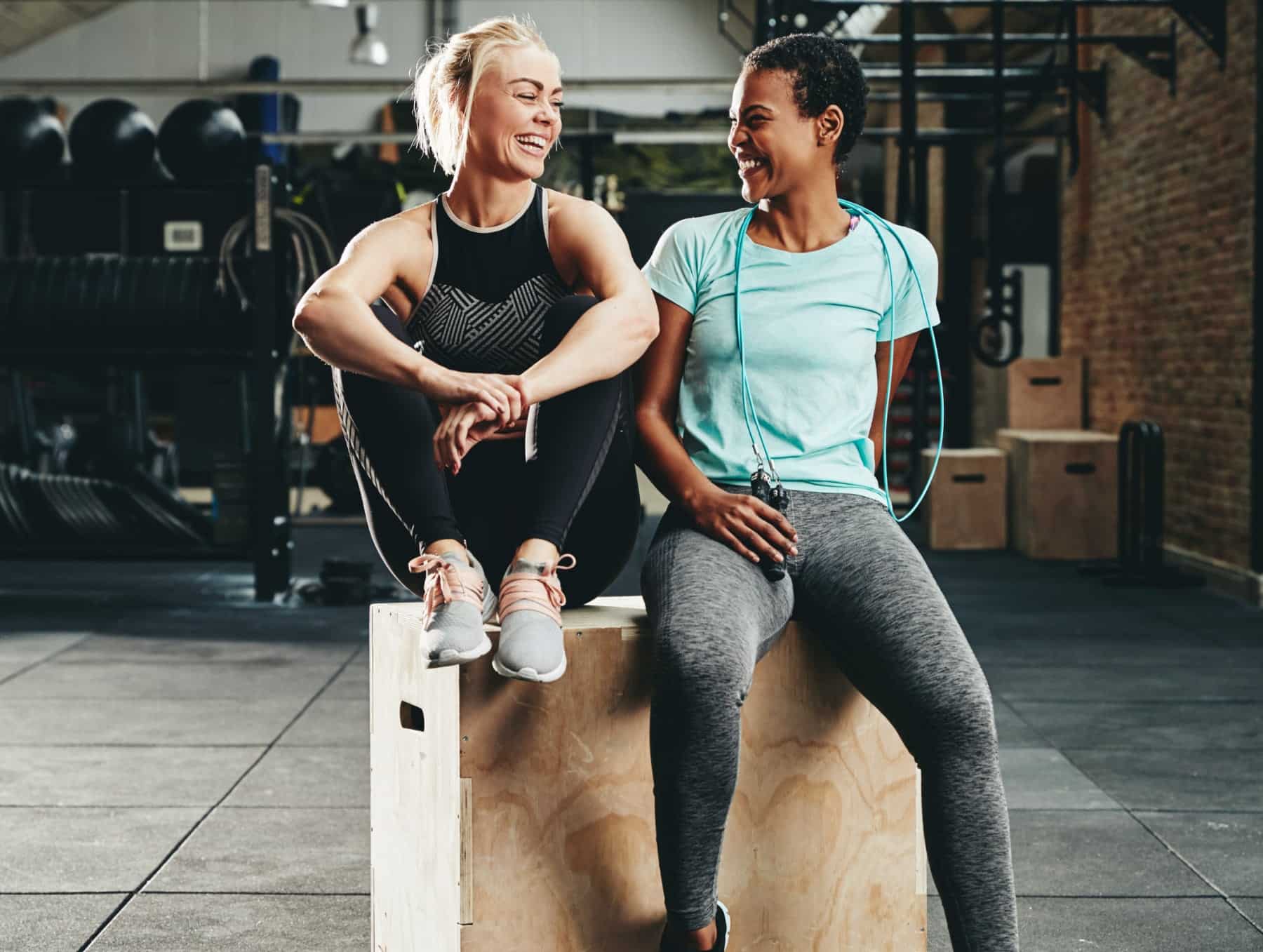Two women over the age of 40 sitting on a box jump in the gym after a workout.