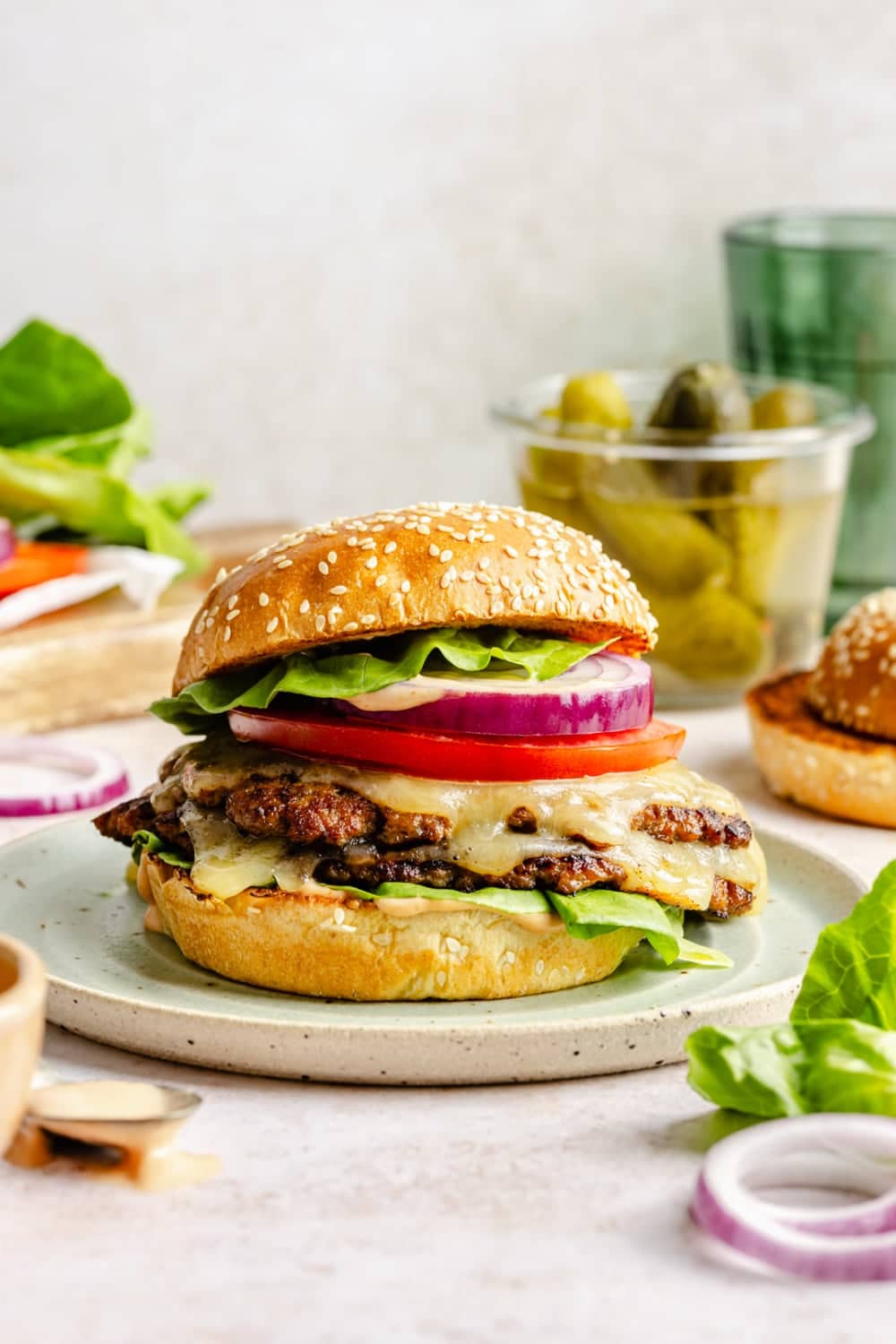 A burger on a plate surrounded by toppings, including lettuce, pickles, tomato, and onion.