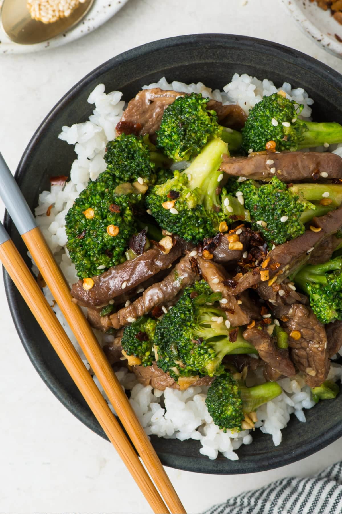 A large bowl with rice and healthy beef and broccoli stir fry on top. It's sprinkled with sesame seeds and red pepper flakes and serve with chop sticks.