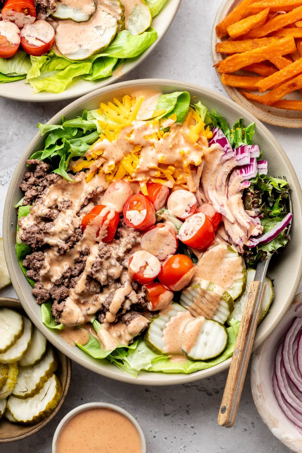 A healthy burger bowl complete with all the ingredients topped with special sauce and served on a table with a side of sweet potato fries.