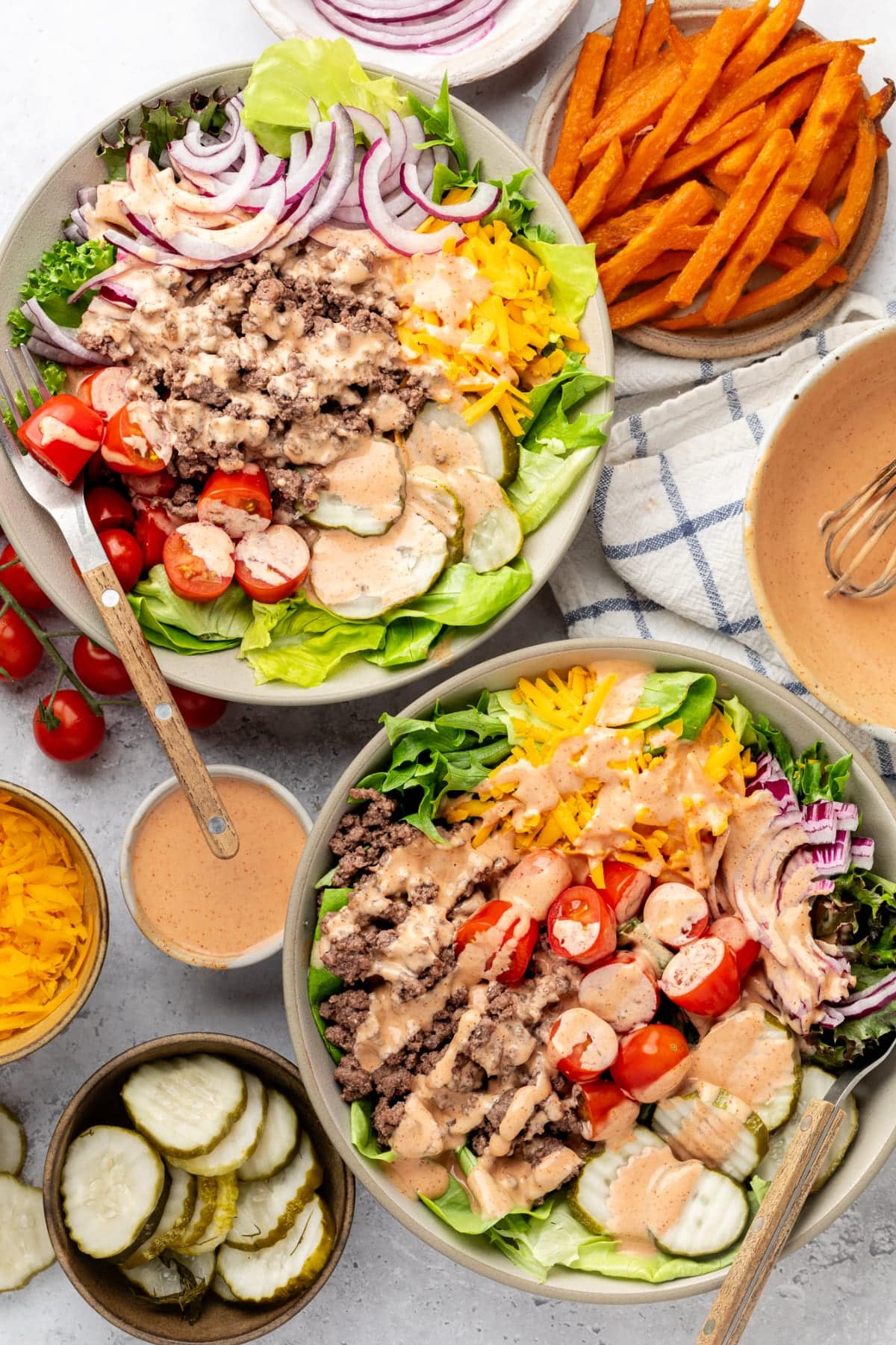 Two healthy burger bowls with special sauce drizzled on top on a table with sweet potato fries, a fork, and a napkin.