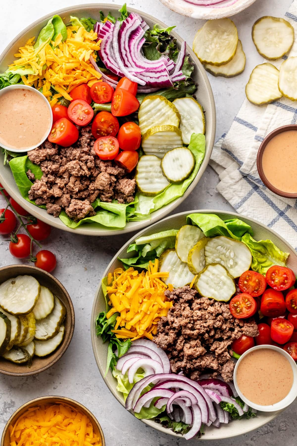 Two bowls side by side on a table surrounded by the special sauce, pickles, and other salad ingredients.
