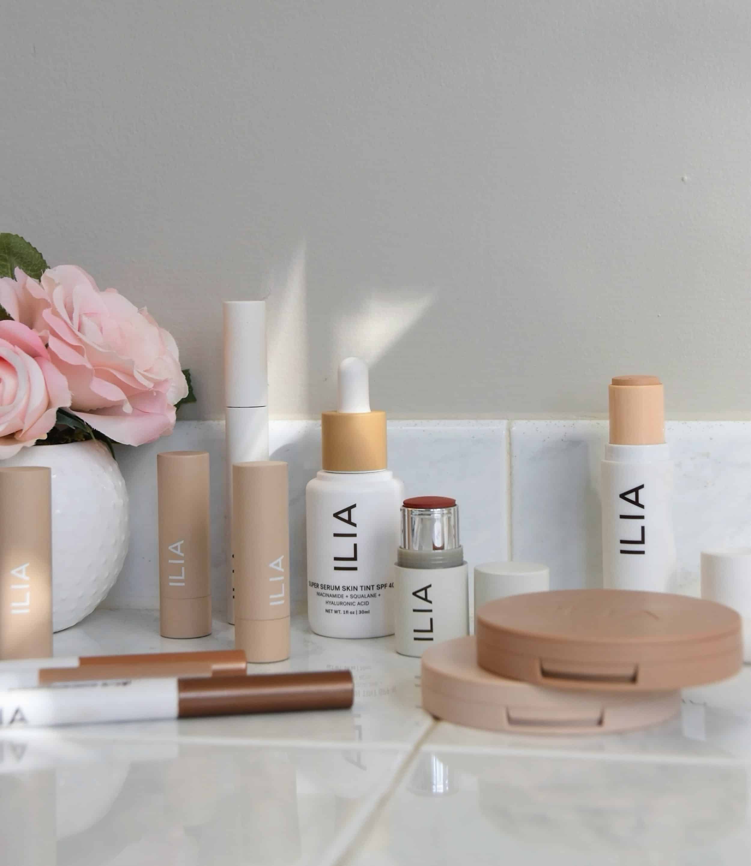 All of the ILIA products in the review on the bathroom counter next to a vase with roses.