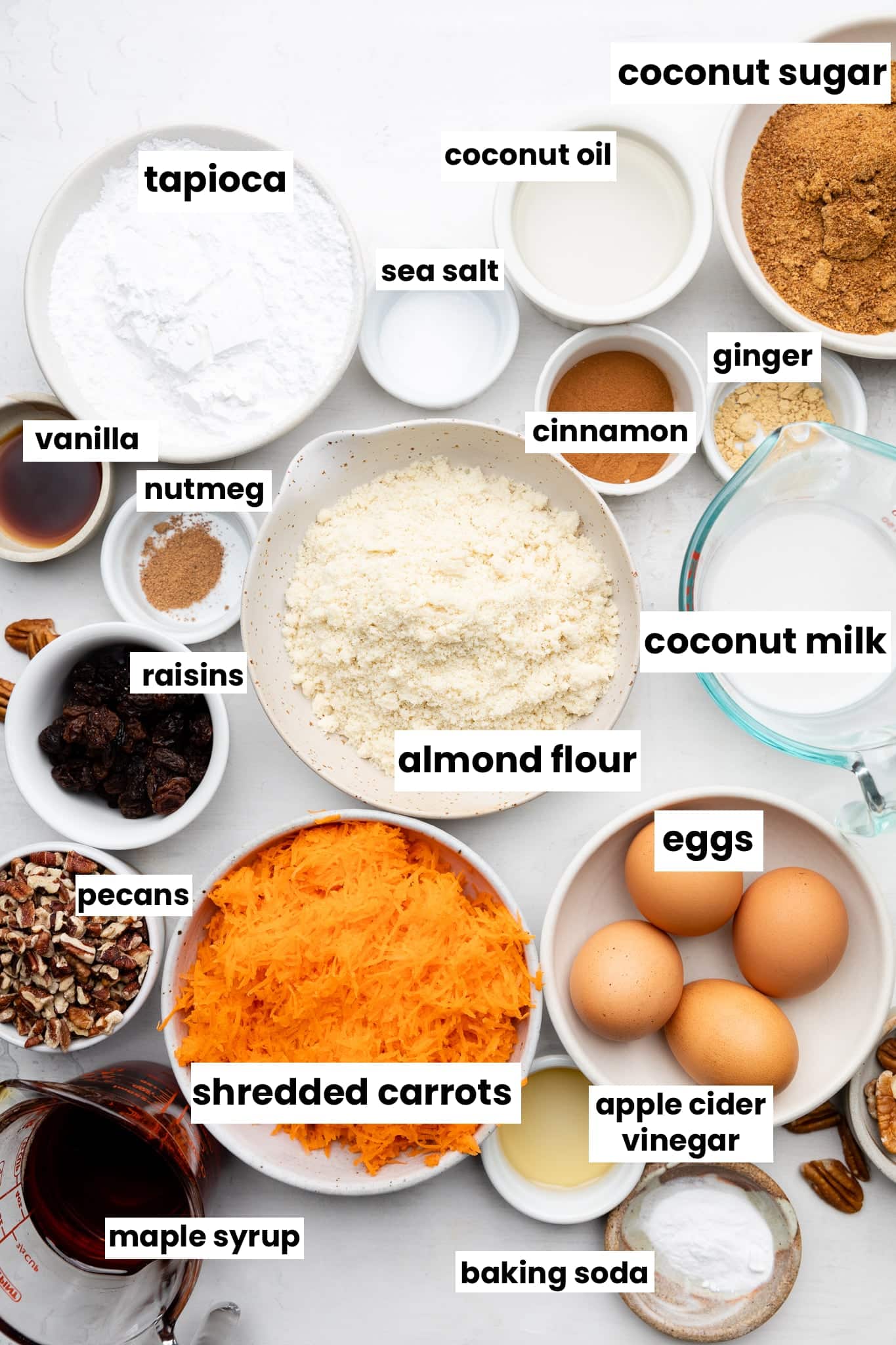 Healthy ingredients in bowls including almond flour, shredded carrots, tapioca, coconut sugar, coconut oil, ginger, cinnamon, vanilla, raisins, coconut milk, pecans, baking soda, eggs, and maple syrup.