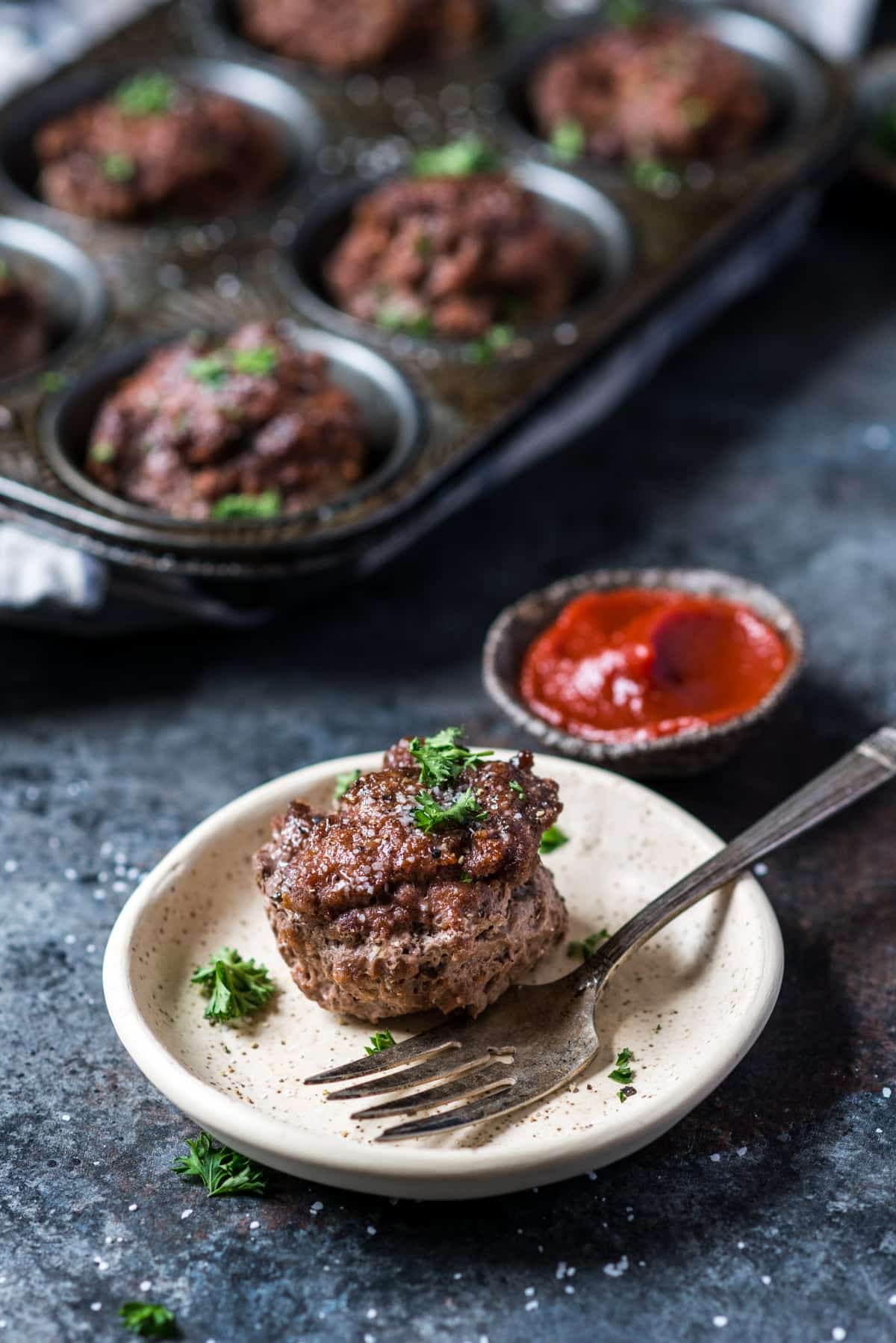 A tin with mini meatloaves and a plat with one mini meatloaf and a fork for eating.