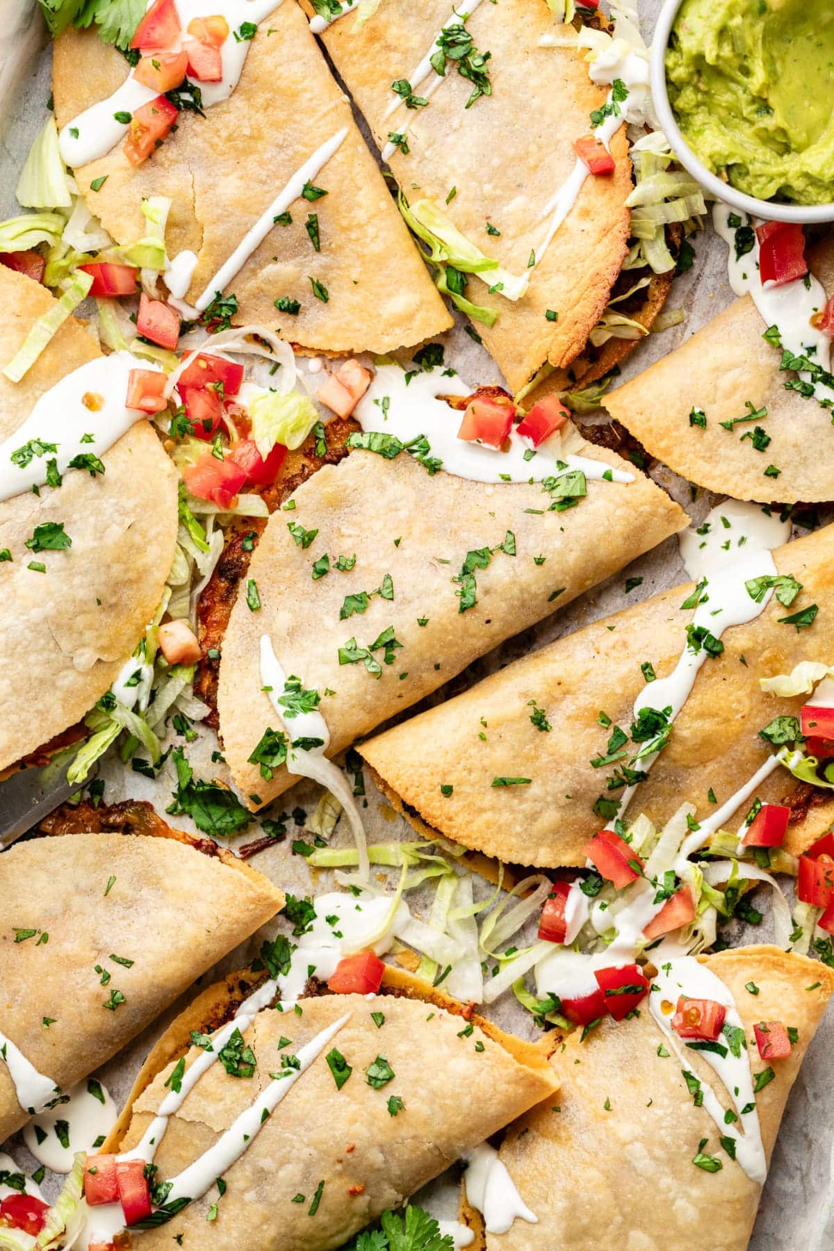 A sheet pan fresh out of the oven with ten crispy baked tacos covered in all the toppings, including guacamole, lettuce, tomatoes, and sour cream.