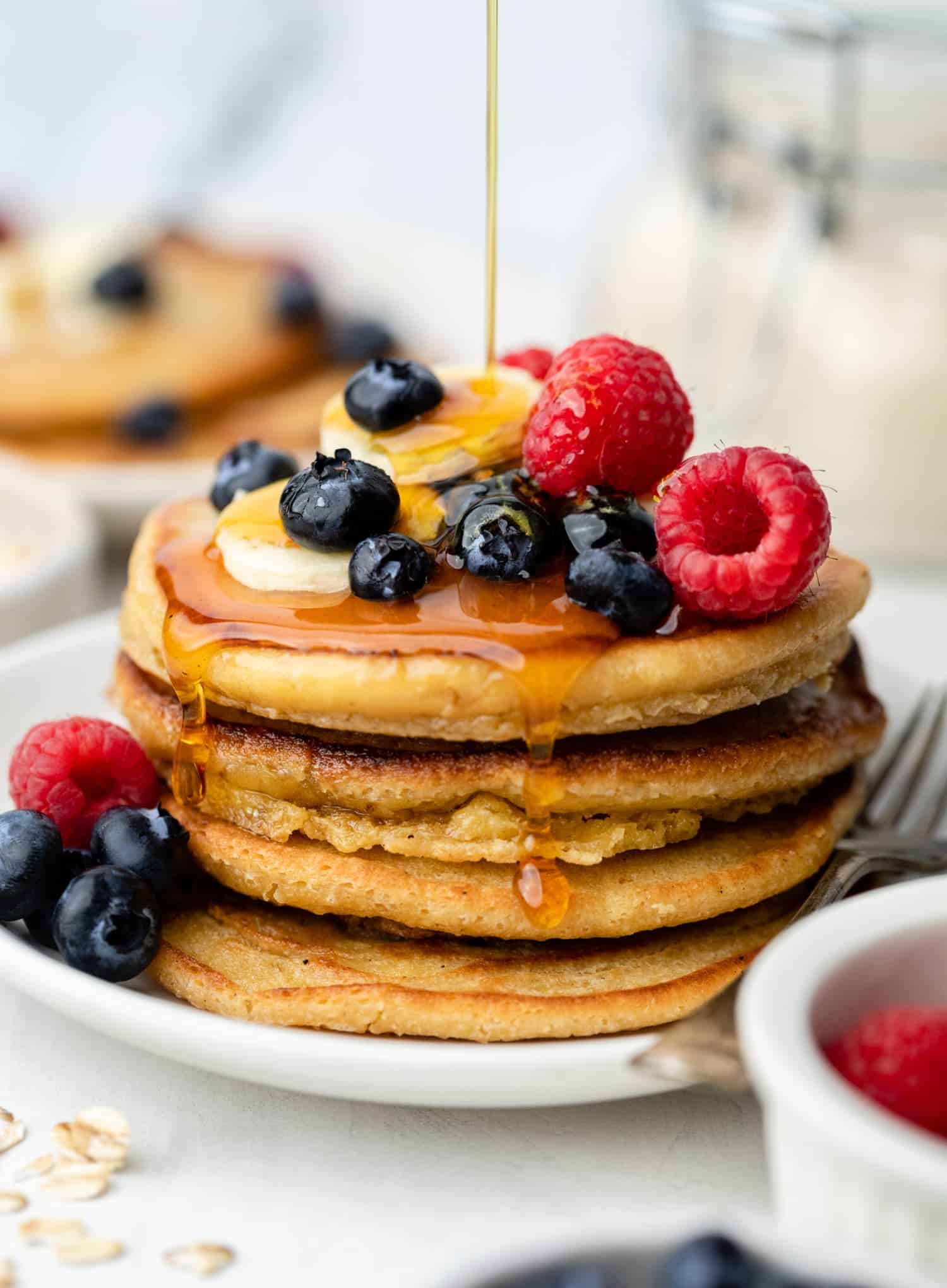 A stack of oat flour pancakes with maple syrup being poured on top. 