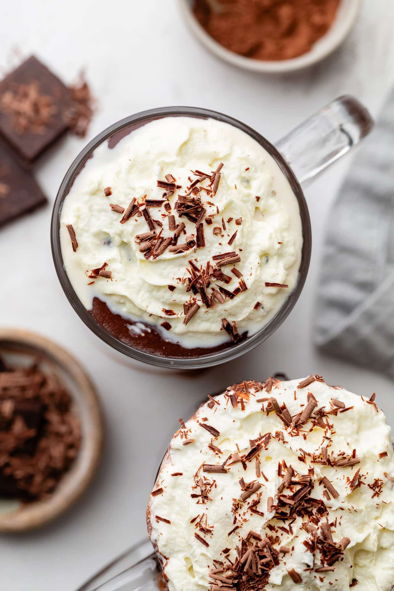 Overhead of two mugs of protein hot chocolate with whipped cream and chocolate shavings on top.