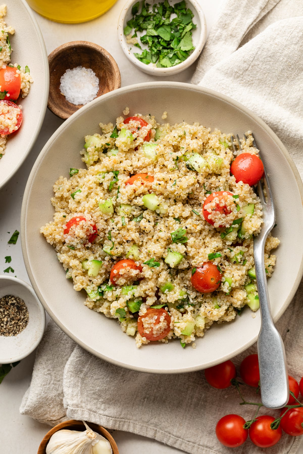 A finished quinoa cucumber tomato salad on a plate with a fork surrounded by a napkin, spices, and tomatoes.