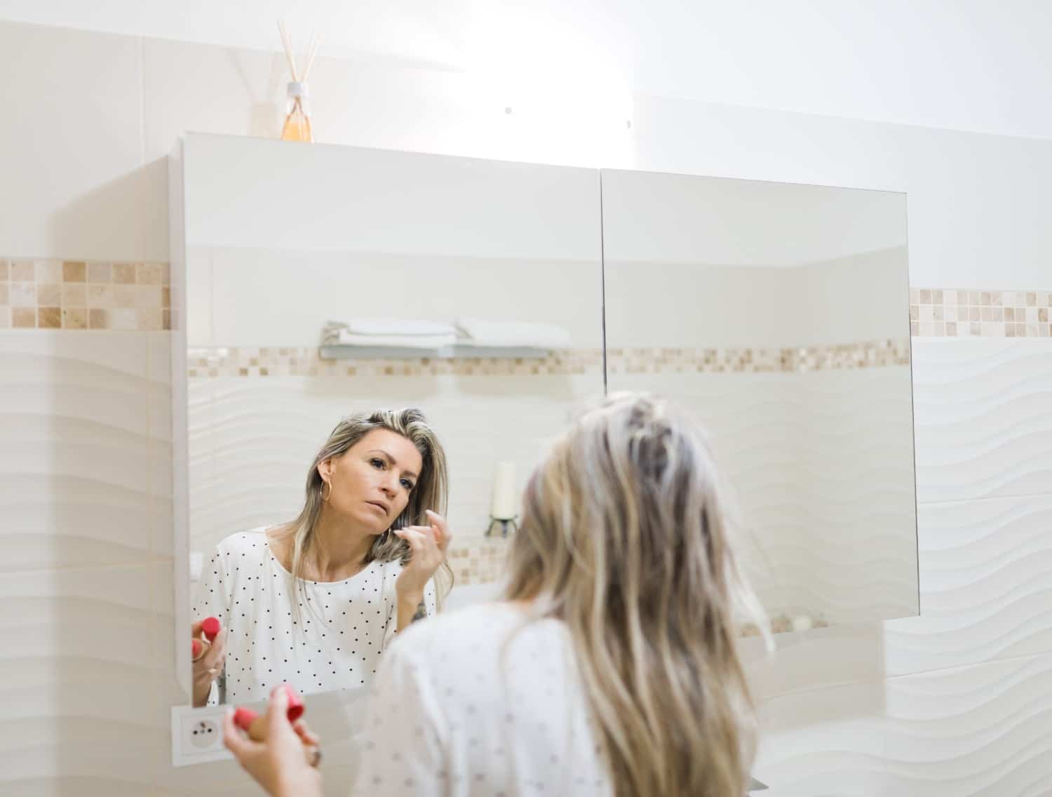 A woman looking in the mirror putting makeup on her hormonal acne. 