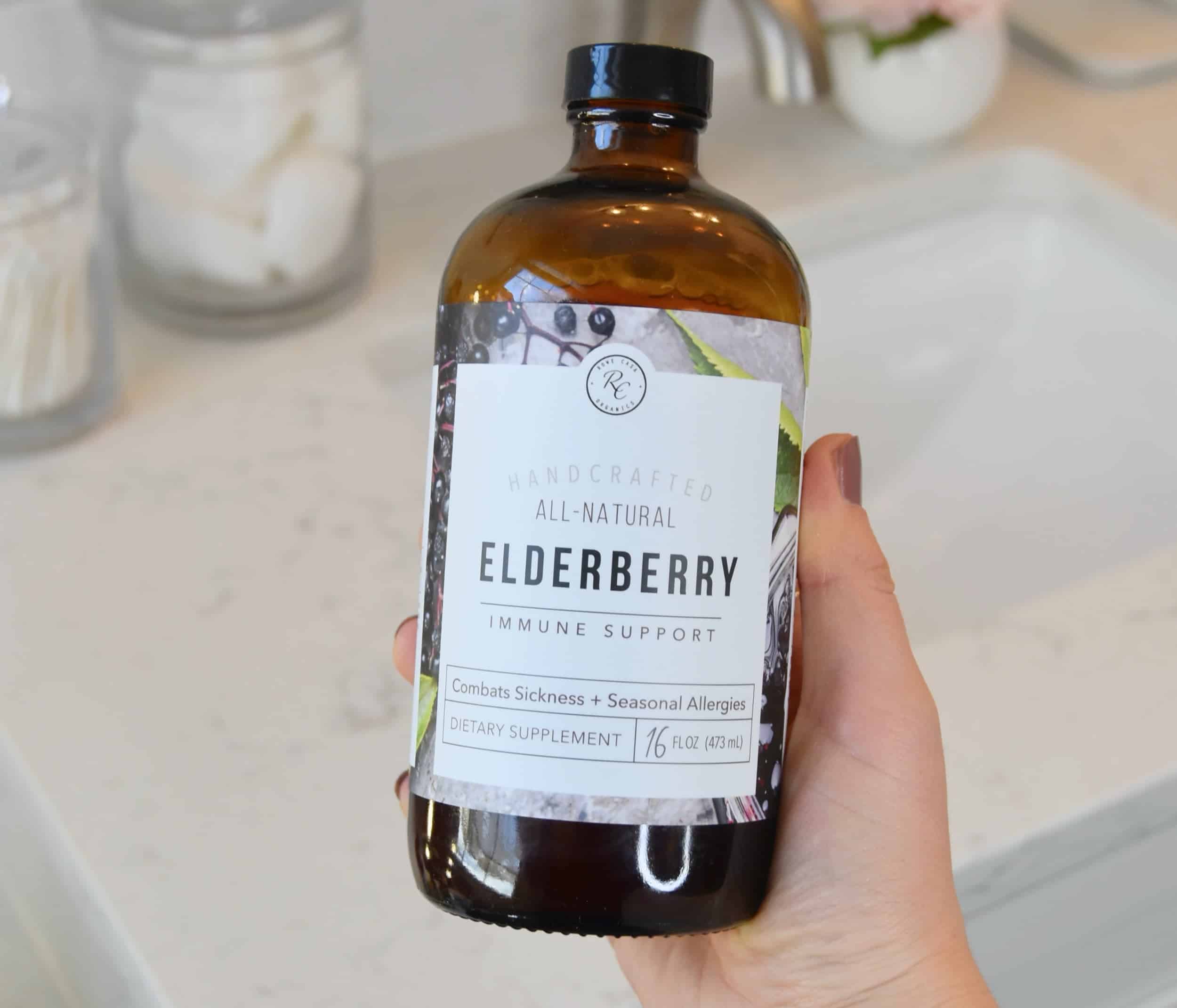 Holding a bottle of Rowe Casa Organics Elderberry Immune Support in front of a marble counter.