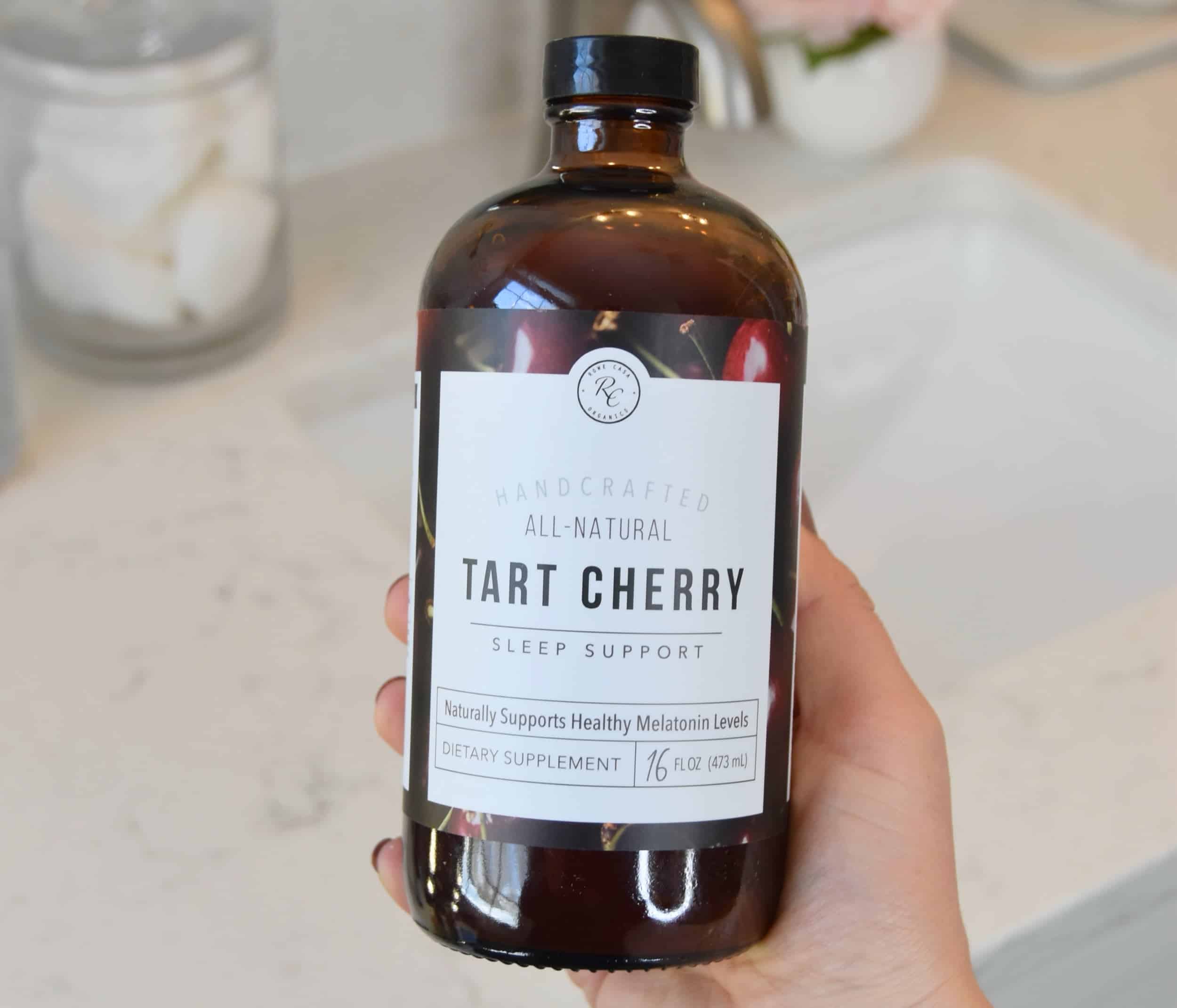 Holding a bottle of the All Natural Tart Cherry Sleep Support in front of a marble counter.