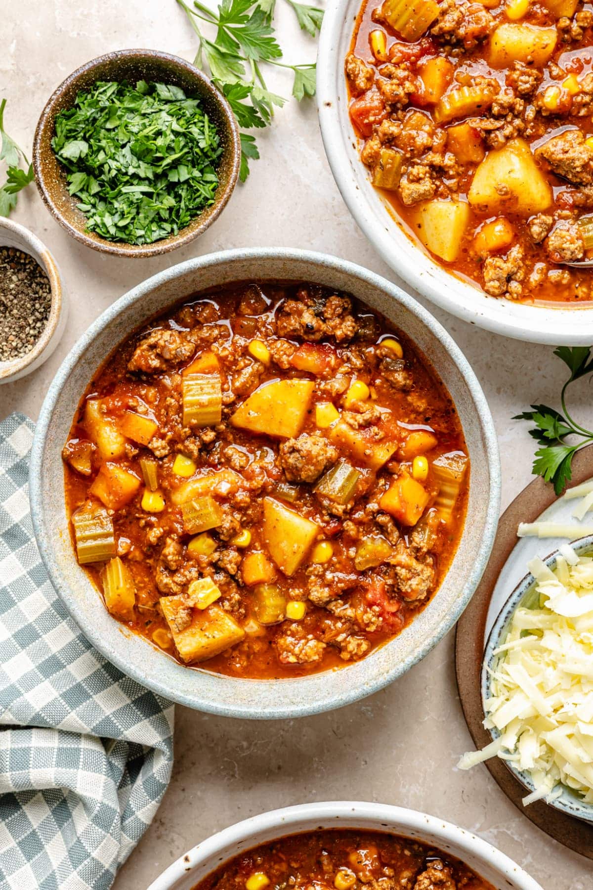 A bowl of slow cooker hamburger soup on a nice dinner table surrounded by a bowl with garnishes, including basil and shredded cheese.