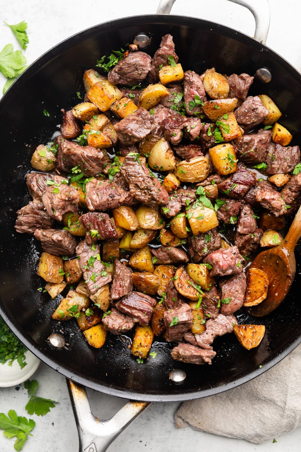 A skillet with finished garlic steak bites and potatoes with a wooden spoon.